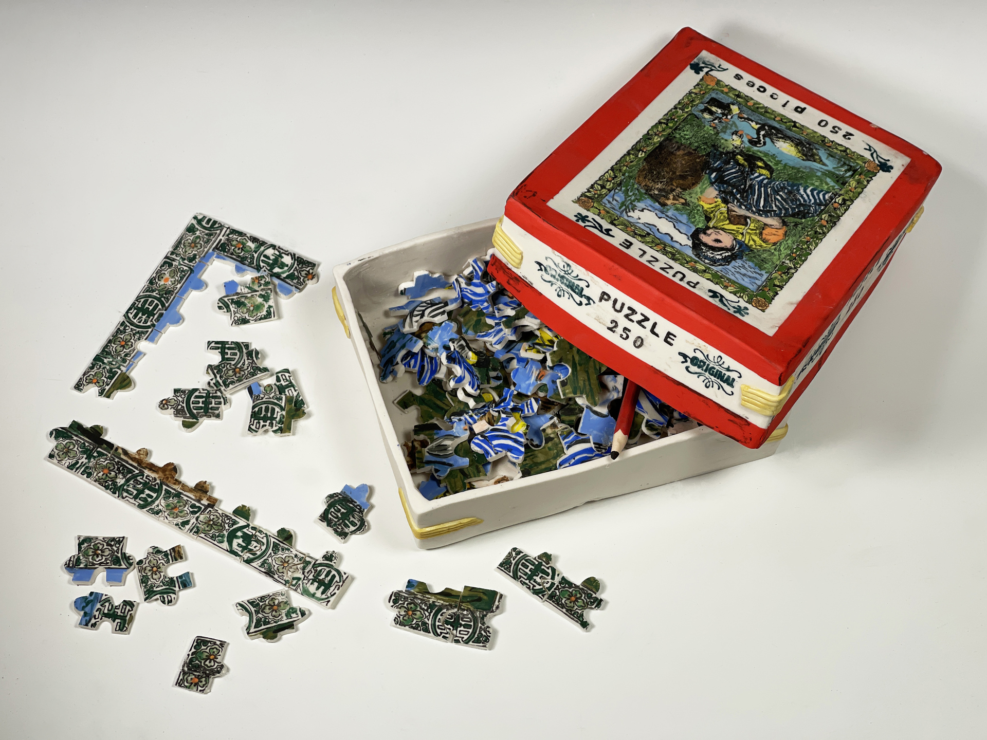 Suzanne Sidebottom used porcelain clay, underglazes  to create a realistic image of a box of puzzle pieces with pieces scattered around it. The box top is a realistic rendering of picture of the finished puzzle.