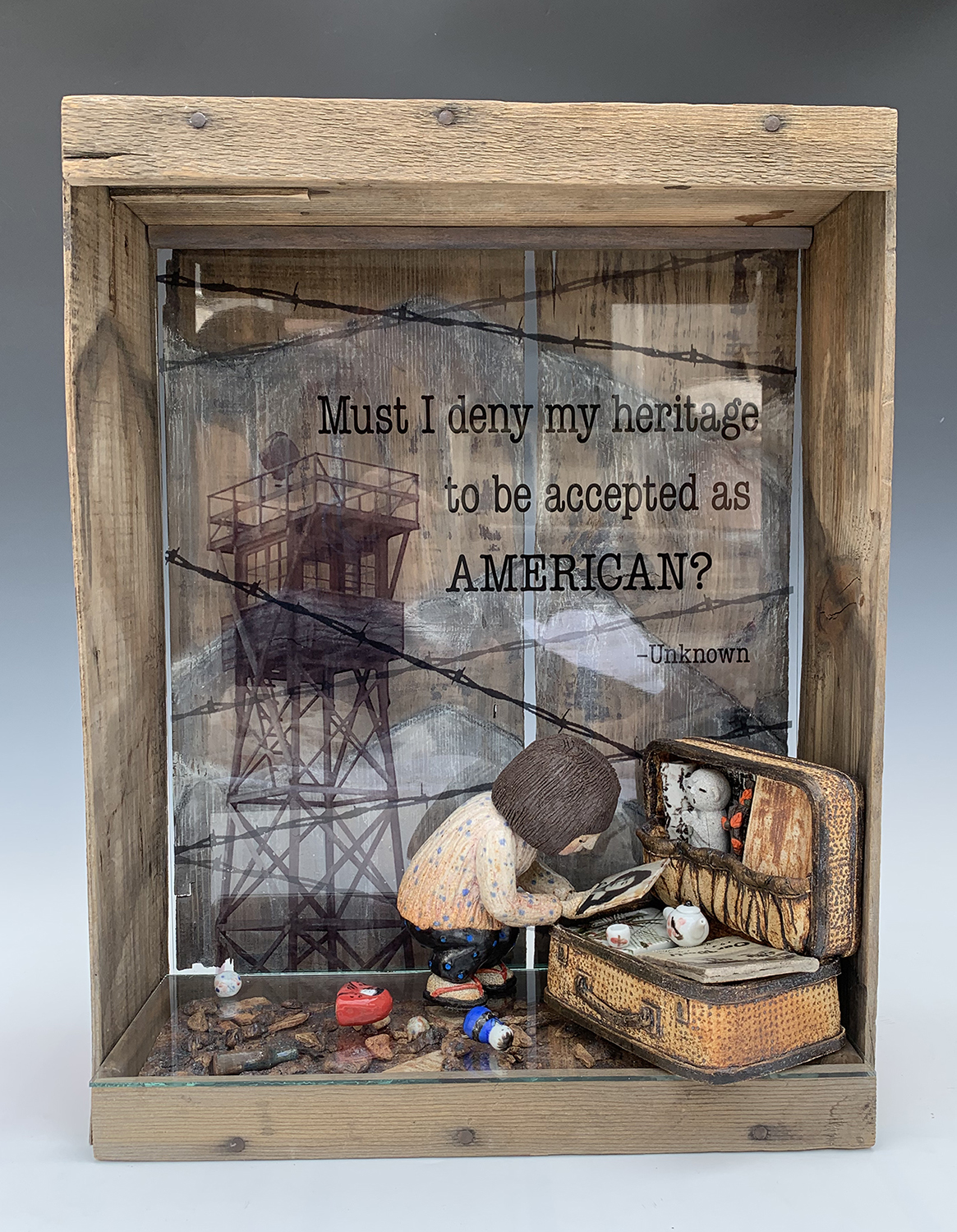 This sculpture by Kathy Yoshihara is a memorial to a relative who was interned with Japanese Americans during World War Two. It contains a figure kneeling over an open suitcase that recounts that the interned could bring only what they could carry. The figure is placed inside a found box, with a computer generated picture of a guard tower and barbed wire.